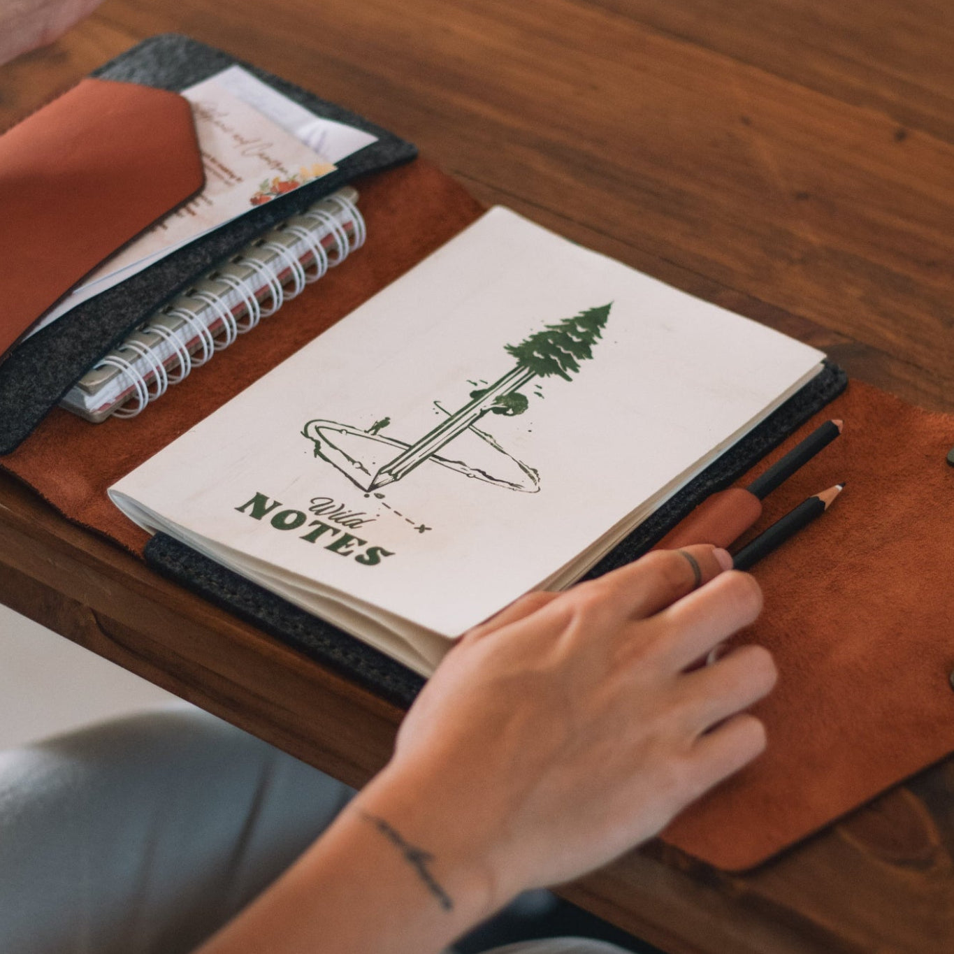 Person holding a mug with a notebook, pen, and pencil on a wooden table