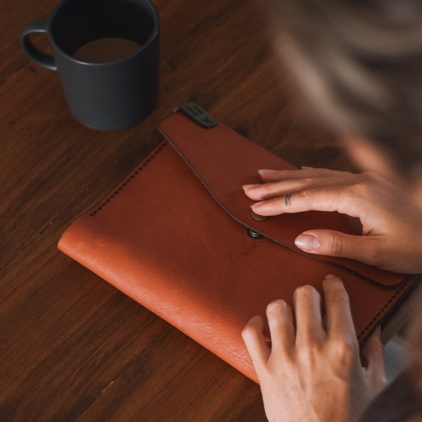 Person holding a brown leather clutch on a wooden table with a black mug in the background